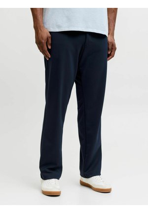 Uomo che indossa pantaloni dritti blu scuro e sneakers bianche, in piedi su uno sfondo chiaro e uniforme.