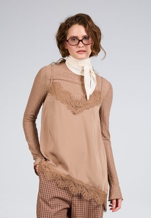 Nainen, jolla on aaltoilevat hiukset ja pyöreät silmälasit, vaaleanbeige pitsireunuksinen tunika pitkähihaisen paidan päällä, kerma huivi ja ruudulliset housut, pukeutuneena yksivärisen taustan edessä poseeraamassa.