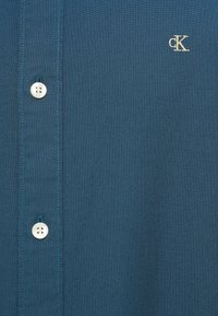 Marškinių tipo palaidinė iš gilaus turkio medvilnės audinio, su baltais mygtukais, kontrastingomis siūlėmis ir auksinės spalvos išsiuvinėtu logotipu kairėje krūtinės pusėje.