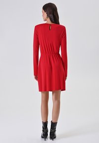 Rode jurk met lange mouwen, een achterkant met een sleutelgat, een getailleerde taille en een zachte textuur, gecombineerd met zwarte laarzen met hak en gestikte accenten.