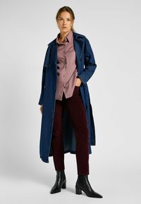 Denim trenchcoat, lång, mörkblå, med dubbelknäppta knappar. Buren över en ljuslila skjorta och burgunderfärgade byxor med svarta klackboots.