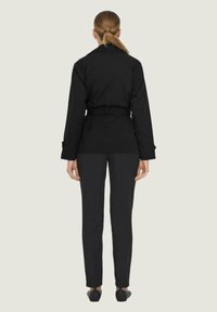 Veste noire à manches longues avec une ceinture à la taille, associée à un pantalon noir slim et des chaussures plates, vue de dos sur un fond neutre.