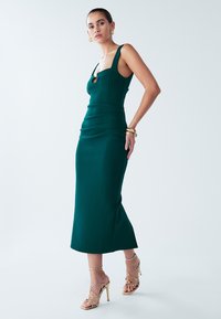 Zielona sukienka bodycon z kwadratowym dekoltem, marszczonym materiałem i rozcięciem po boku. Połączona ze złotymi sandałami na wysokim obcasie z paskami.