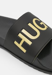 Schwarze Badesandale mit breitem Riemen, der große goldene Glitzer-"FUG"-Buchstaben zeigt, strukturiertem Fußbett und Gummisohle.