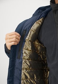 Marineblaue Jacke mit Reißverschluss, die ein glänzendes, goldenes, gestepptes Innenfutter und eine weiche schwarze Außenseite zeigt. Entworfen für Wärme und Stil.