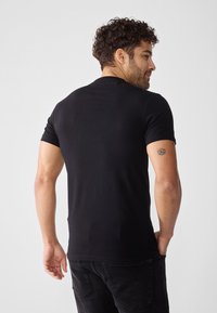 Svart kortermet skjorte med en tettsittende design og myk tekstur, brukt med mørke jeans. Har en synlig tatovering på armen.