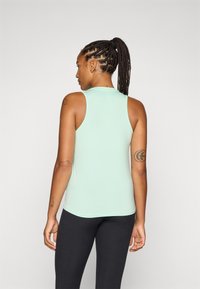 Een persoon met gevlochten haar staat met de rug naar de kijker, gekleed in een lichtgroene tanktop en zwarte legging.