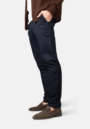 Hombre con pantalones ajustados azul marino, mocasines marrones de ante y un cárdigan marrón, con la mano en el bolsillo, de pie contra un fondo blanco.