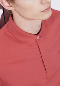 Piké i korallfärgad bomullstyg med en texturerad yta, med spetsig krage, en enkel knappar placket och diskret logotypdetalj.