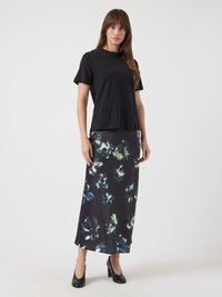 Ženska stoji v črni majici s kratkimi rokavi, črni cvetlični midi krilu in črnih visokih petah, na beli ozadju.