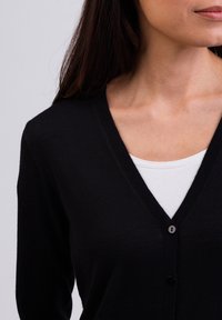 Černý cardigan s výstřihem do V, který má zapínání na knoflíky a ribbovaný povrch. Pod ním je vidět bílá košile s kulatým výstřihem.