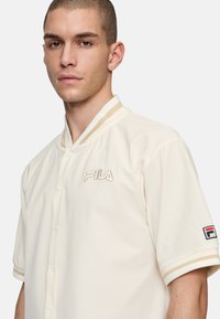 Camicia a maniche corte di colore crema con tessuto rigatino, colletto e polsini avorio con righe dorate, caratterizzata da un logo FILA ricamato.