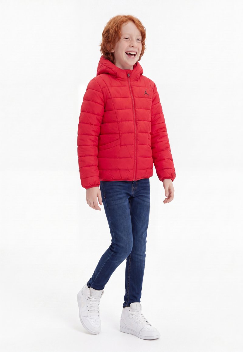 Enfant aux cheveux roux portant une doudoune rouge, un jean bleu et des baskets blanches, souriant et marchant sur un fond blanc.