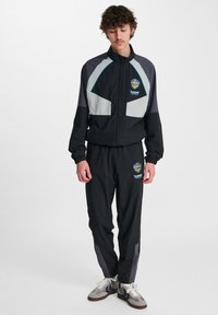 Svart träningsdräkt med gråa och blå accenter. Jackan har dragkedjestängning och logotyp. Byxorna har resårmidja och smalnande ben.