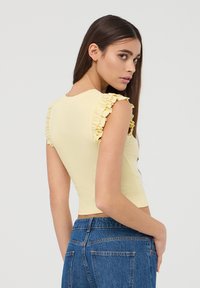 Giovane donna con lunghi capelli scuri che indossa un crop top giallo pallido con maniche a ruches e jeans a vita alta blu, che guarda oltre la spalla.