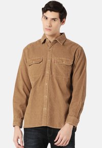 Brown corduroy shirt with long sleeves, button front, two chest pockets, and a small logo on the left pocket. Textured fabric design.