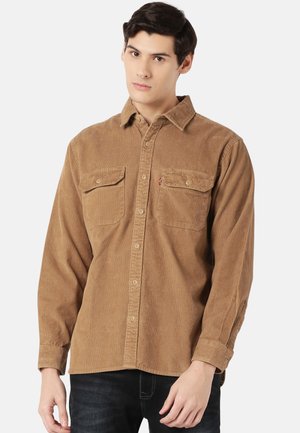 Camisa de pana marrón con mangas largas, botones en la parte delantera, dos bolsillos en el pecho y un pequeño logo en el bolsillo izquierdo. Diseño de tela texturizada.
