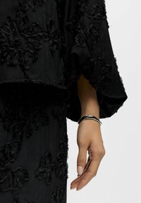 Ranką puošia sidabrinis apyrankė, iš dalies uždengta juodu audiniu su tekstūra ir gėlėmis ant rankovės bei drabužio.