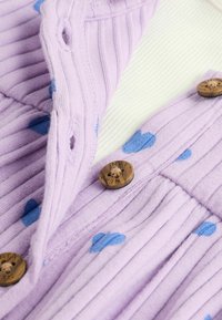 Violeta ribota auduma ar zilu sirdsapziņu rakstu, kurā ir koka pogas. Dizains ietver tekstūras un kontrastējošas krāsas vizuālai pievilcībai.