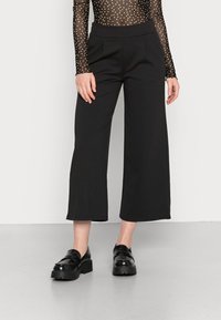 Zwarte brede broek met een elastische tailleband, met zijzakken en een gladde, matte textuur. Draagt met zwarte plateauschoenen.