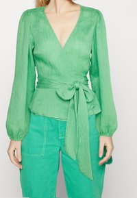 Zelený top s hlubokým výstřihem do V, dlouhými nabíranými rukávy, strukturovanou látkou a detaily vázání v oblasti pasu, kombinovaný se shodnými zelenými kalhotami.
