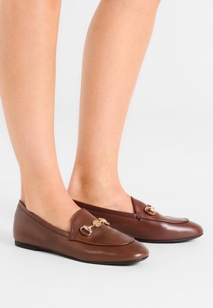 Mocassins en cuir marron avec un détail en métal doré portés pieds nus, vus de profil sur un fond blanc.
