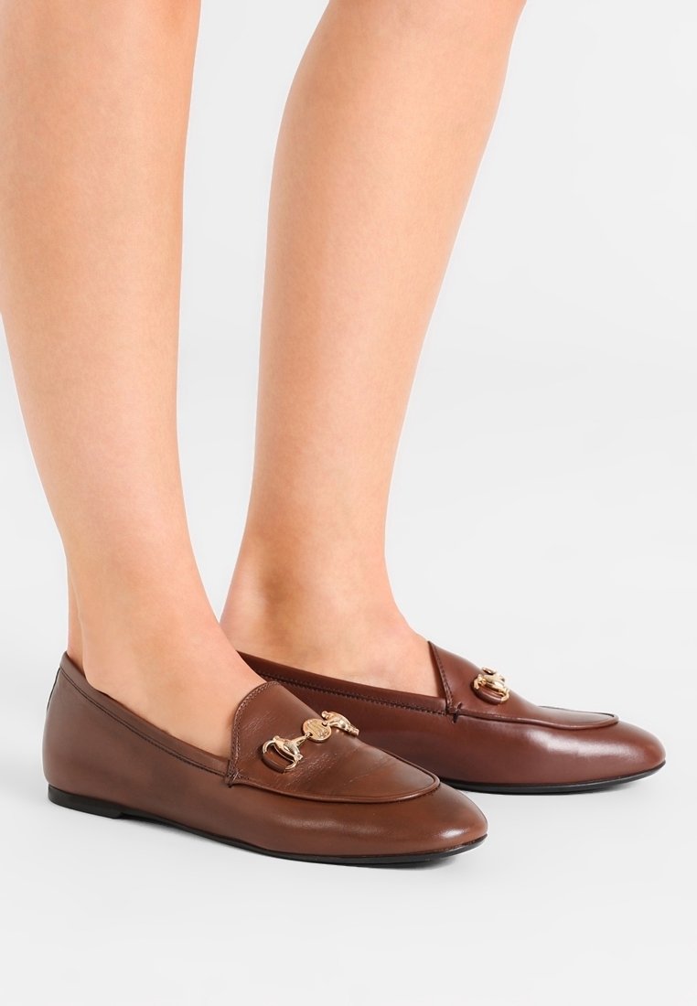 Mocassins en cuir marron avec un détail en métal doré portés pieds nus, vus de profil sur un fond blanc.