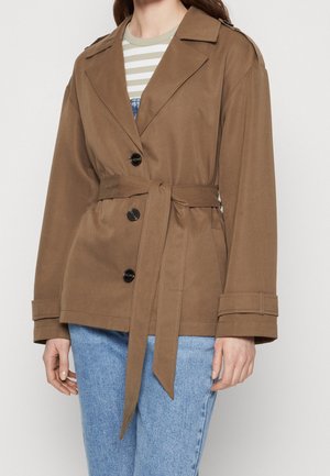 Vrouw draagt een bruine jas met ceintuur en zwarte knopen over een gestreept shirt en lichtblauwe spijkerbroek, staand tegen een effen achtergrond.