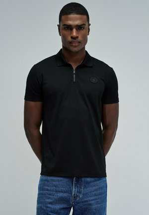 Jeune homme aux cheveux courts portant un polo noir zippé et un jean bleu, debout devant un fond uni gris clair.