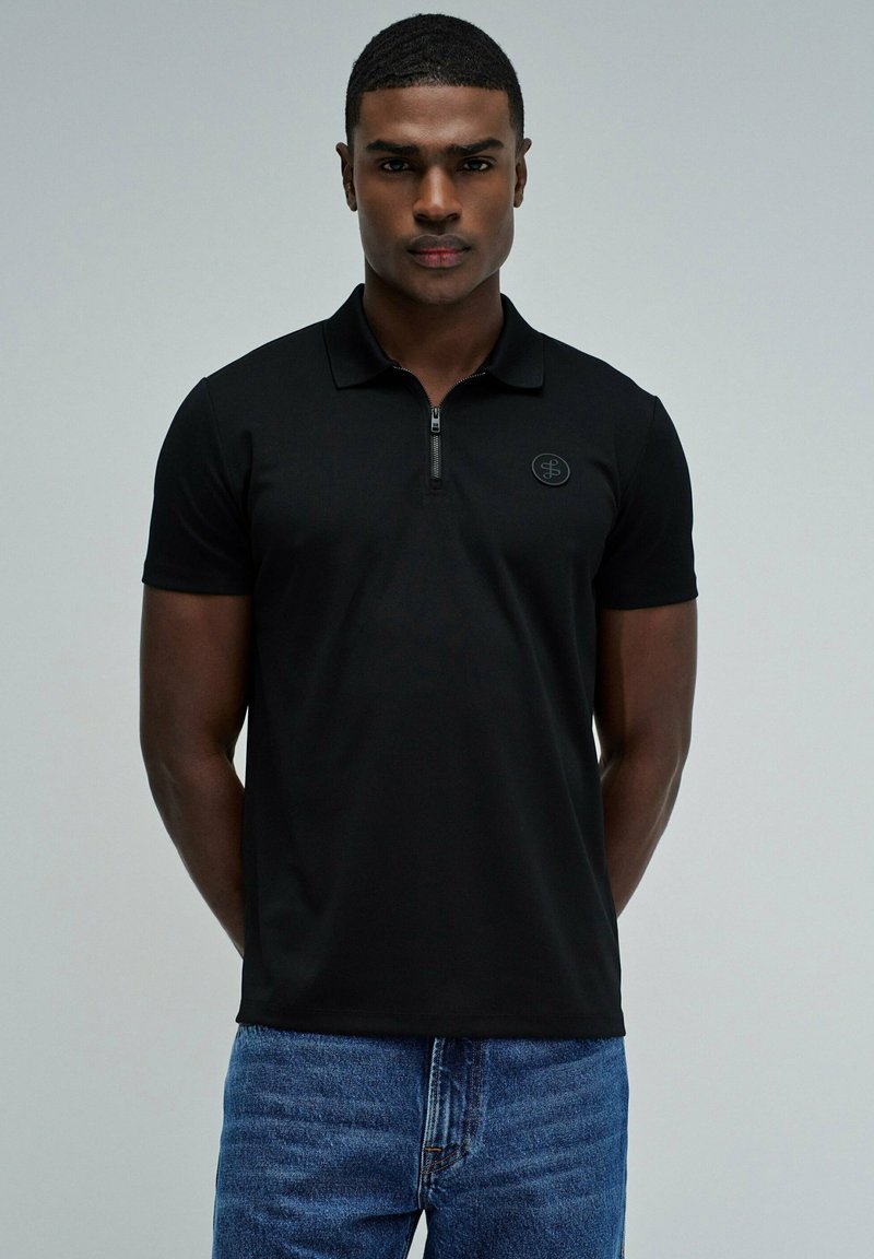 Joven con el pelo corto que lleva una camiseta tipo polo negra con cremallera y vaqueros azules, de pie frente a un fondo gris claro liso.