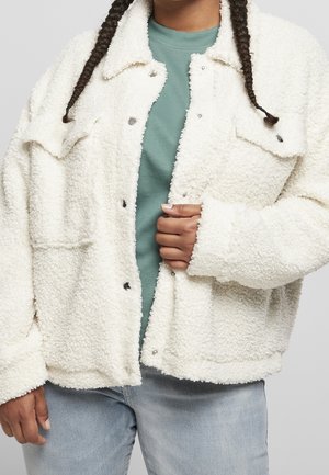 Person med to fletninger iført en hvid tekstureret skjortejakke over en blågrøn skjorte og lyseblå jeans, der holder jakken lukket.