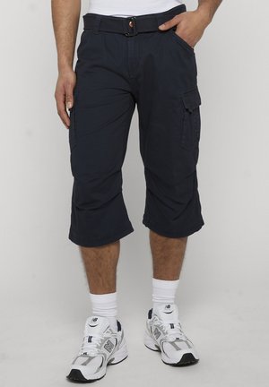 Hombre con pantalones capri cargo negros con bolsillos laterales, calcetines blancos y zapatillas blancas New Balance, de pie contra un fondo liso.