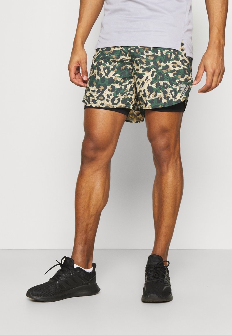 Homme portant un short de sport camouflage vert et beige avec une doublure intérieure noire et des chaussures de course noires, debout devant un fond uni.
