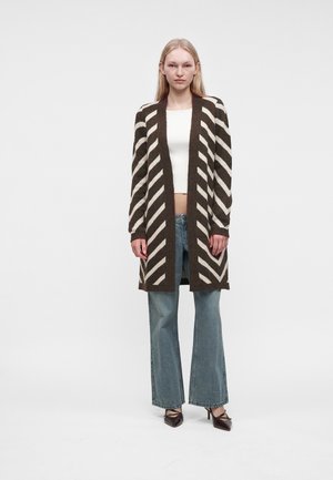 Жена, облечена с дълъг кафяво-бял райе кардиган, бял къс топ, сини дънки и кафяви обувки на ток, стои пред обикновен фон.