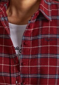 Rode geruite flanellen shirt met blauwe en witte lijnen, met knoopsluitingen en geborduurd logo. Witte T-shirt zichtbaar eronder.