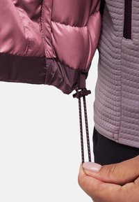 Hand die trekkoord van een mauve puffer jacket aanpassen naast de ribbelige mavelkleurige mouw van een jas op een effen achtergrond.