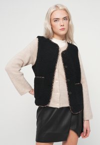 Μαύρο γιλέκο από φλις με λεοπάρ σχέδιο στις άκρες, διαθέτει κουμπιά μπροστά και δύο τσέπες, φορεμένο πάνω από ένα μπεζ πουλόβερ και μαύρη δερμάτινη φούστα.