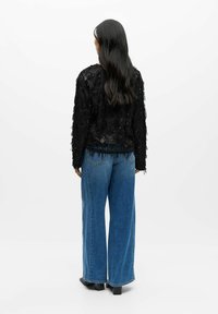 Žena s dlouhými tmavými vlasy oblečená v černém strukturovaném topu s dlouhým rukávem a modrých širokých kalhotách, stojící zády k bílému pozadí.