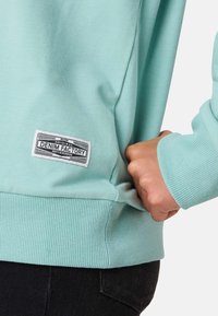 Felpa turchese chiaro con polsini e orlo a coste. Presenta un'etichetta che dice "DENIM FACTORY" sul lato, con una texture liscia e morbida.