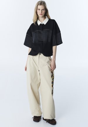 Młoda kobieta stoi, ubrana w czarną koszulkę z krótkim rękawem i białym kołnierzykiem oraz szerokie beżowe spodnie z bocznym lampasem w panterkę.