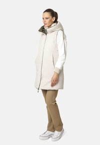 Chaleco acolchado sin mangas color beige claro con capucha, que presenta una textura acolchada y un cierre frontal. Lleva encima una camiseta blanca de manga larga y pantalones beige.