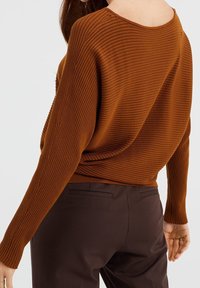 Vrouw die een roestkleurige ribgebreide trui met een open rug en donkerbruine, op maat gemaakte broek draagt, gezien van achteren tegen een effen achtergrond.