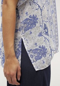 Blusa ligera y estampada con diseños florales azules sobre una base blanca, manga corta y una abertura lateral para mayor movilidad.