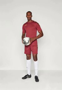 Maillot de sport rouge à manches courtes, shorts assortis, chaussures de football noires et chaussettes blanches, tenant un ballon de soccer à motifs.