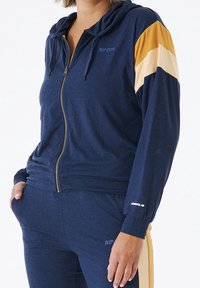 Veste à zip bleu marine avec des accents en or et crème rayés sur les manches, en tissu doux, et avec une capuche à cordon. Le pantalon présente un design assorti.