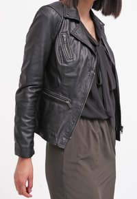 Veste motard en cuir noir avec poches zippées, coupe ajustée et détails matelassés aux épaules. Elle est associée à un haut noir et une jupe olive.