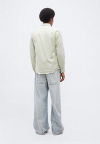 Άνδρας με αφροχτένισμα, φορώντας ένα ανοιχτό πράσινο πουκάμισο με ρίγες και άνετο ανοιχτό μπλε τζιν, στέκεται γυρισμένος πλάτη σε απλό φόντο.