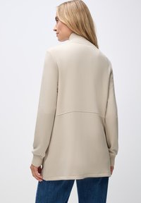Gilet long beige à manches longues avec un col et des poignets côtelés, présentant une coupe décontractée et une texture de tissu lisse. La vue de dos met en avant les détails de couture.