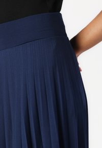 Marineblaue, plissierte Bluse mit einem hohen Bund, aus strukturiertem Stoff gefertigt. Verfügt über vertikale Falten für zusätzlichen Schwung und Detail.