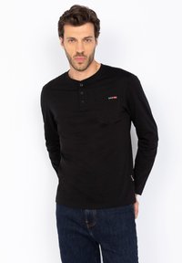 Maglietta henley nera a maniche lunghe con scollatura a girocollo, patta con tre bottoni e taschino sul petto. Realizzata in tessuto morbido con una vestibilità ampia.